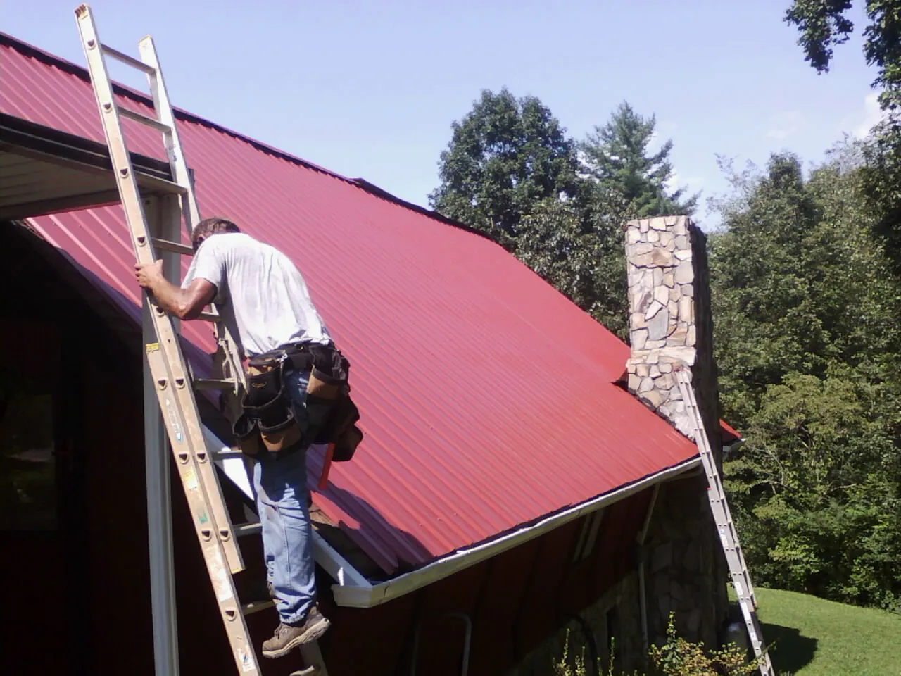 Roofer on a ladder beside a red metal roof and stone chimney — Soffit & Fascia Repair by Roofing Brothers in Erlanger